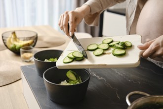 A person slices cucumbers on a wooden cutting board, adding them to two black bowls filled with