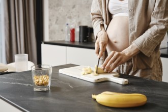 A pregnant woman slices a banana while standing at a countertop. A glass of nuts and a banana are