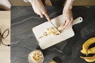 A person slices fresh bananas on a white cutting board in a well-lit kitchen. The countertop
