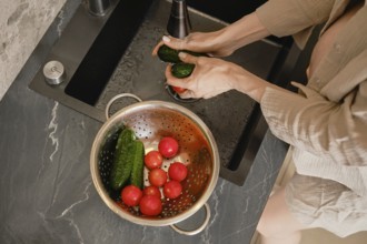 Hands scrub cucumbers under running water at a sleek kitchen sink. A colander nearby holds ripe