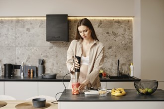 A pregnant woman stands in a contemporary kitchen using a handheld blender to prepare a fruit