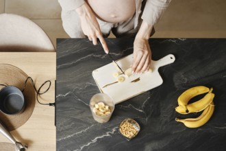 A person with a visible baby bump is cutting bananas on a wooden cutting board in a modern kitchen.