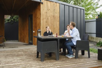 A family gather around a black table on a wooden deck of tiny house, sharing drinks and laughter in