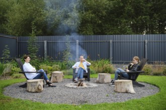A family relax around a circular fire pit, seated on rustic chairs during a weekend. Smoke drifts