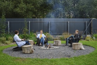 A family enjoy a relaxing evening around a fire pit in a backyard. They are sitting on wooden