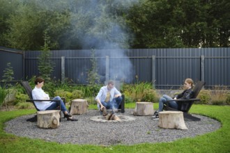 A family sits comfortably on wooden chairs, gathering around a fire pit in a spacious garden. Smoke
