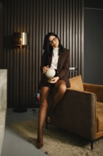 A businesswoman sits on a brown armchair, holding a globe while wearing a tailored suit. Concept of