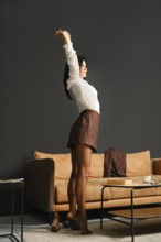 A slender woman dressed in a white blouse and a brown skirt stands on tiptoe and stretching arms in