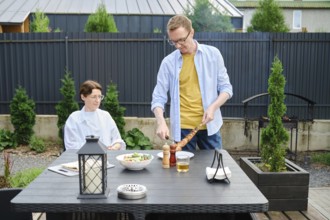 A man is serving grilled meat to a woman seated at a dining table in a beautiful backyard. The