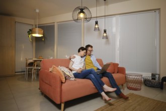A husband embraces his wife while sitting on a couch in a modern living room. They are watching a