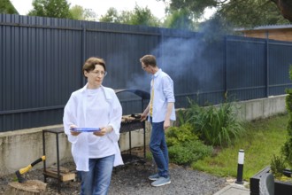 Two people are preparing a barbecue outdoors. A woman holds a plate while walking away, and a man