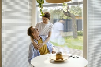 A couple shares a joyful interaction at a modern kitchen table. The woman embraces the man from