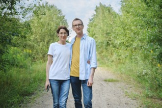 A middle-aged couple stands closely together on a natural path surrounded by lush greenery. They
