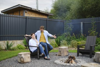 A couple spends a relaxed evening outdoors near a fire pit in their backyard. They share playful