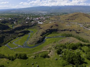 River flowing through a hilly landscape with a city in the background under a blue sky, aerial