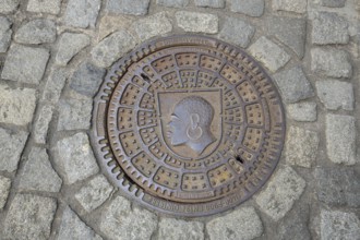 Round manhole cover with city coat of arms, inscription, cobblestones, floor, head, African,