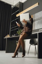 A woman in a stylish outfit is leaning against the edge of her desk in an office. She holds a