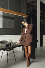 In an office, a woman wearing a brown blazer and skirt is engaged on a phone call while reviewing