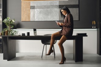 Serious business woman in a brown suit sits on a desk, focused on her smartphone. She checks her