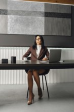 A slender woman with glasses and a stylish suit sits at an office desk with opened laptop. She