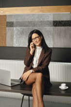 Positive businesswoman engages in a phone conversation while seated at a table. A laptop and a