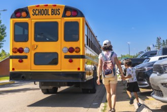 Mother holding son's hand walking towards a school bus on the first day back to school,