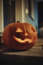 A carved, glowing pumpkin with a smiling face stands on a wall in front of a house at night,