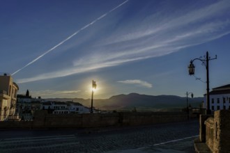 Sun rising behind mountains, illuminating street lamps on cobblestone bridge at dawn in ronda,