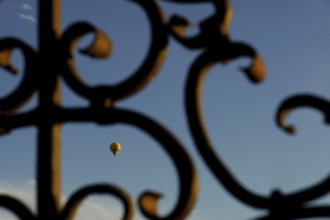 Yellow hot air balloon flying against a blue sky, seen through an ornate metal gate in ronda,