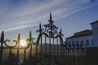 Sun shining through the ornate wrought iron gate at sunrise in ronda, spain, with mountains and
