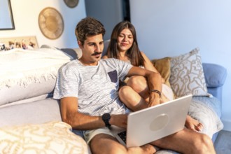 Young couple sitting comfortably on sofa browsing internet on laptop, enjoying time together at