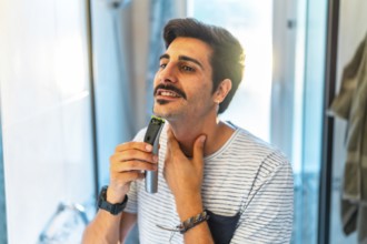 Man trimming his beard using an electric razor, taking care of his appearance in the bathroom
