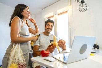 Young couple smiling and enjoying bananas while working on a laptop in a bright, modern home
