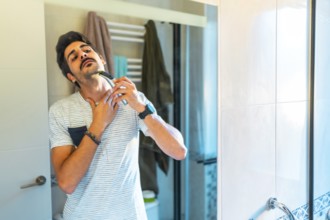 Young man carefully shaving his neck with an electric razor, looking in the bathroom mirror during