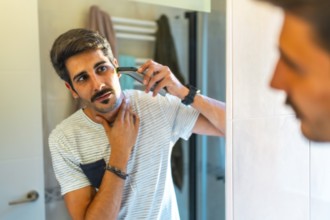 Concentrated young man trimming his beard with electric razor in bathroom mirror, morning routine,