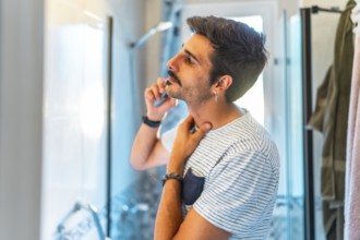 Young man trimming his mustache with an electric razor and touching his neck while looking in the