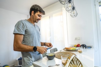 Young man spreading butter on fresh bread while enjoying breakfast in a bright, modern kitchen