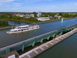 A passenger ship sails on a canal over a large bridge, surrounded by city buildings and nature,