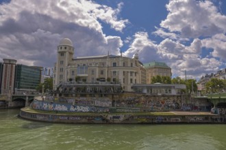 A historic building by the river with graffiti, under a clear sky, surrounded by urban surroundings