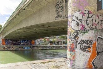 Under a bridge on the river with graffiti-covered columns and riverbank surroundings, Am Donaukanal