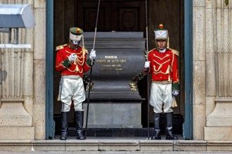 Two uniformed soldiers in red jackets stand guard in front of a marble building, Two soldiers guard