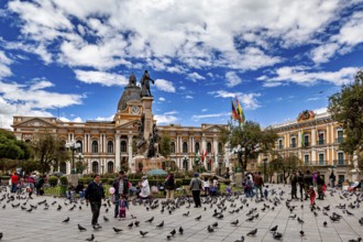 Main square full of people and pigeons in front of a large building with a statue under a blue sky,