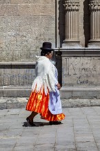 Woman in traditional dress with orange skirt walking along a historical wall, Traditionally dressed