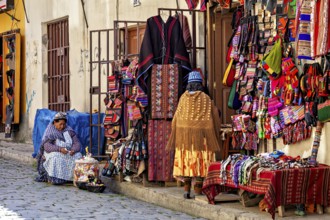 A woman sits in front of a market stall selling a variety of colourful textiles and traditional
