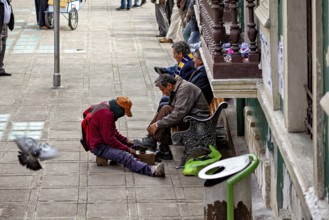 A shoeshine boy shines shoes on a street while passers-by and a pigeon are visible, shoeshine boy