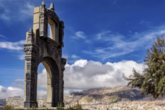 A stone gate with ornate decorations in front of a sprawling cityscape and a blue sky, The gate at
