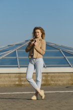 A young woman stands outdoors, wearing a light jacket and jeans. She holds her jacket gently while