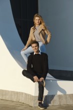 A young man sits on a low wall with his leg bent while a woman stands above him, both smiling. The