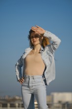 A woman stands outdoors on a sunny day, shielding her eyes from the sunlight. She wears a light