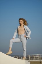 A young woman stands on a ledge outdoors, wearing light denim and looking directly at the camera.